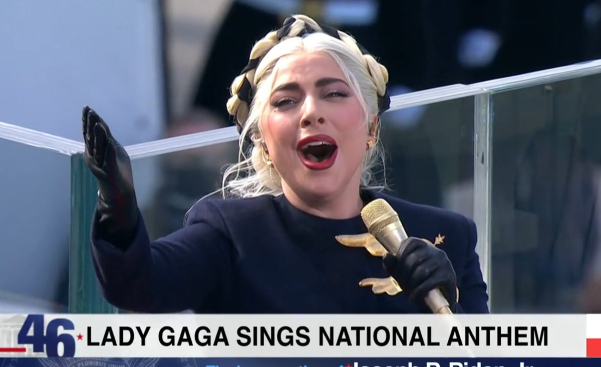 Lady Ga Ga singing the national anthem wearing a blue and gold suit. She is holding a gold microphone. There is a gold bird on her dress.