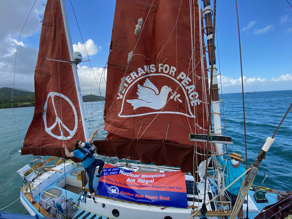 Honololu, the Golden Rule sailboat at sea. A woman holds the sails, while standing by a sign saying nuclear weapons are illegal. photo:  Connie Durant Golden Rule Veterans for Peace project. 