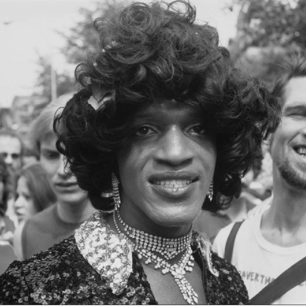 Black and white portrait of Marsha P. Johnson, a Black drag queen. The photo is taken from the shoulders up, she is wearing a curled bob, blouse and diamond jewelry, and smiling toward the camera. Photo credit is given to Dr. Ron Simmons. 