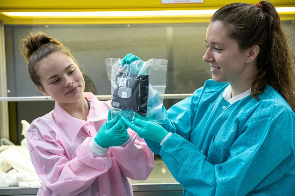 Emily Kennebeck and another woman in clean room suits holding a scientific instrument in a plastic bag
