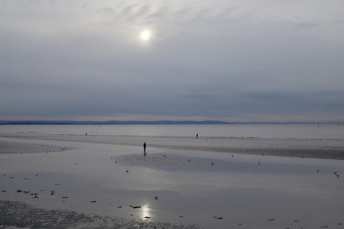 The last day of January, a cold grey afternoon at Crosby beach. The pale white disc of a watery winter sun adds a silvery sparkle to pools of water left by the outgoing tide as it lowers into the approaching cloud mass over the snow covered hills of North Wales. <a href="/IronMenCrosby/">IronMenCrosby 🌤</a>