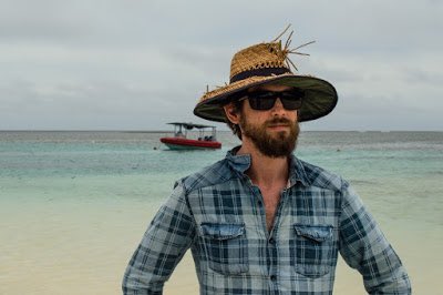 Dan stands in front of the ocean wearing a straw sun hat and sun glasses. There is an small boat in the background.