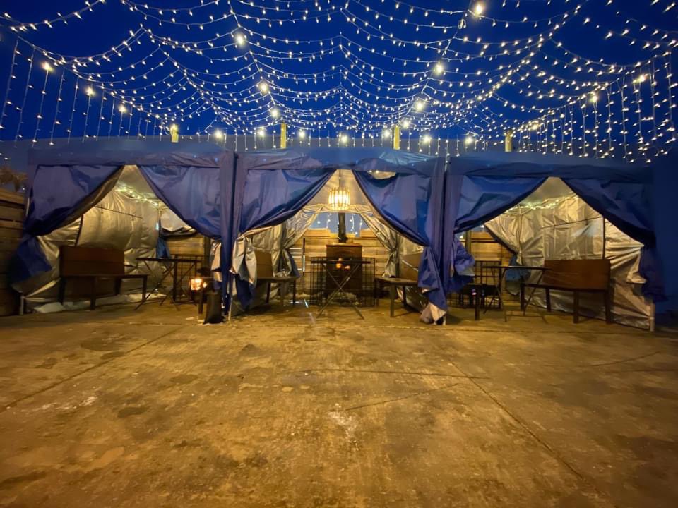 Three outdoor tents, with benches, wood stoves, and tables. Overhead fairy light, illuminate an outdoor patio.