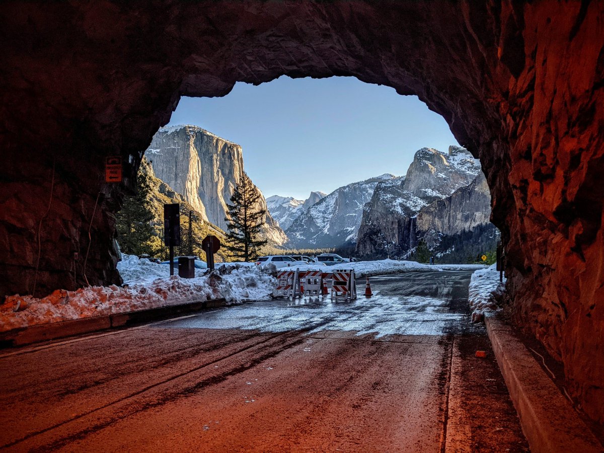 Looking out to Yosemite Valley from just inside the Wawona Tunnel. Soft red lights along tunnel walls, snow covers the edge of the road and mountain tops in the distance.