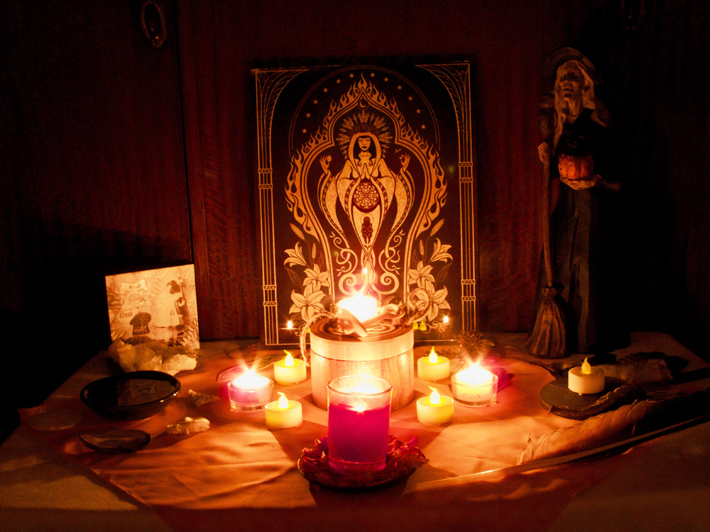 photo of tea-light candles  on an altar. Various objects on a small table are bathed in orange and red light; four tea light candle are white-hot, aglow with flame, with a thin line of light emanating from each candle.