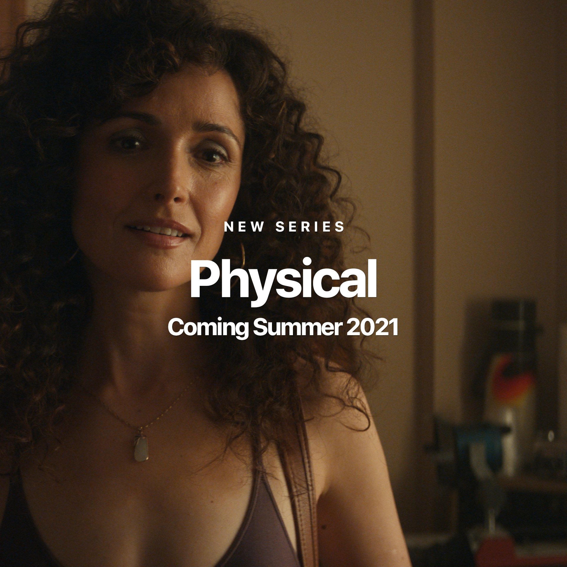 A woman with very curly hair looks into the camera as if she’s about to say something in a still pulled from the Apple Original series, Physical.