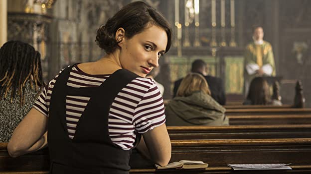 Phoebe Waller-Bridge kneeling in a church pew looking over her shoulder, wearing a black and white shirt
