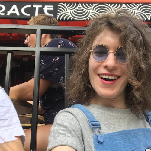 Le visuel est une photo d'une jeune femme. Celle-ci a les cheveux bouclés, porte des lunettes de soleil rondes et sourit. 