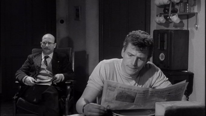 Black and white still from sixties film featuring Johnny Briggs in the foreground reading a newspaper.