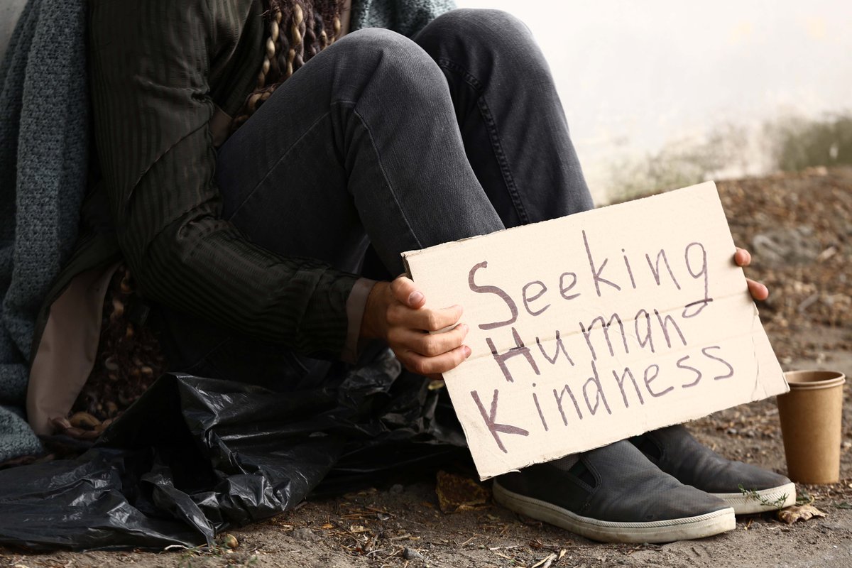 Person sitting on ground holding cardboard sign that says "seeking human kindness."