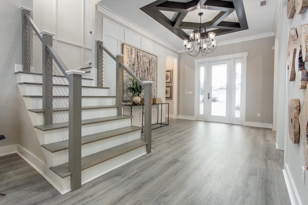 A wide foyer with a dramatic stained beam ceiling detail, set to a tension cable stairwell railing backdrop makes for a stunning entry into this home. #modelhome #foyerdesign   #stainedbeamdetail  #coastaldesign   #watersoundorigins #modelmerchandising #masterpiecedesigngroup