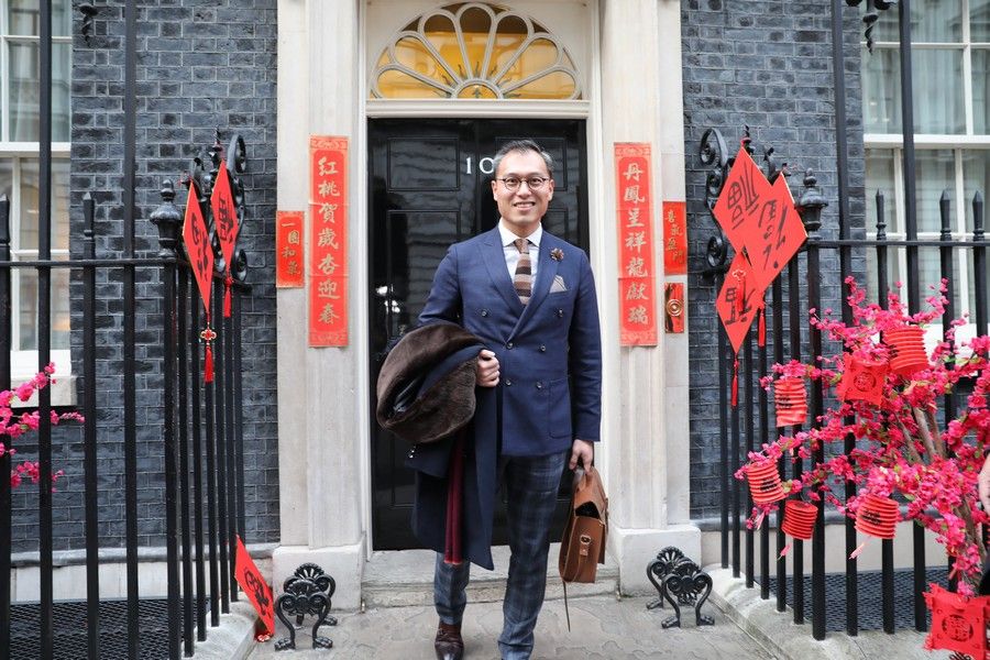 photo of a smiling Chinese man in a suit outside Number 10 Downing Street. He is holding a briefcase in one hand and had his coat over his other arm. There are red decorations on the walls and railings with Chinese characters on them. 