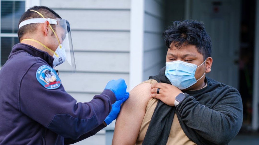 Photo depicting a masked man raising his sleeve to receive a COVID-19 vaccine.