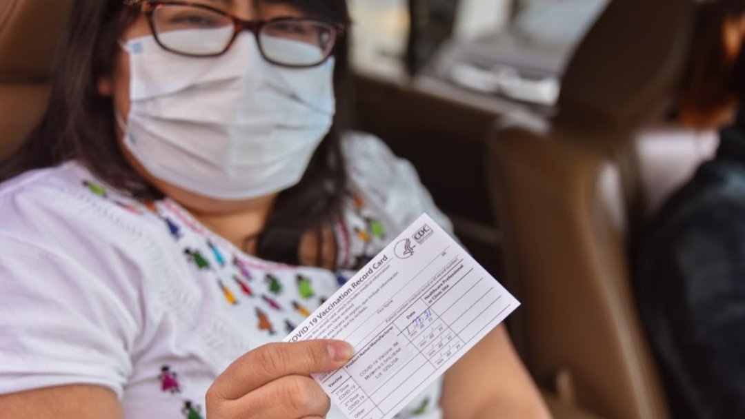 Photo depicting a masked woman in a car holding up her COVID-19 vaccination card.