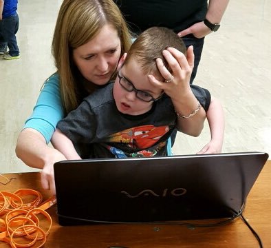 A mom in a blue T-shirt helps a small child in a black T-shirt use a laptop set up with eye gaze equipment to play games