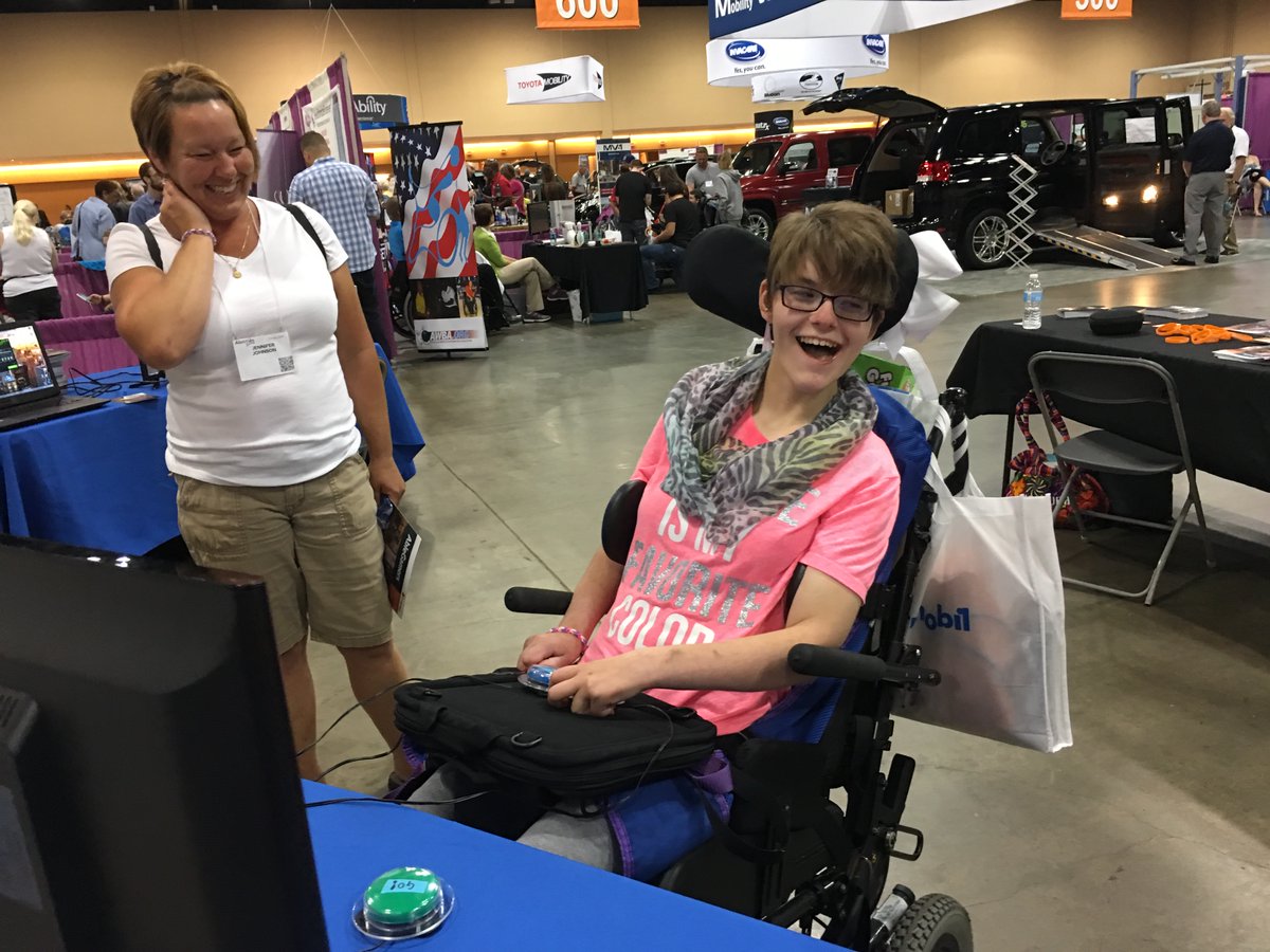 A woman in a white T-shirt looks on at a very happy woman in a pink T-shirt playing video games in a wheelchair for the first time