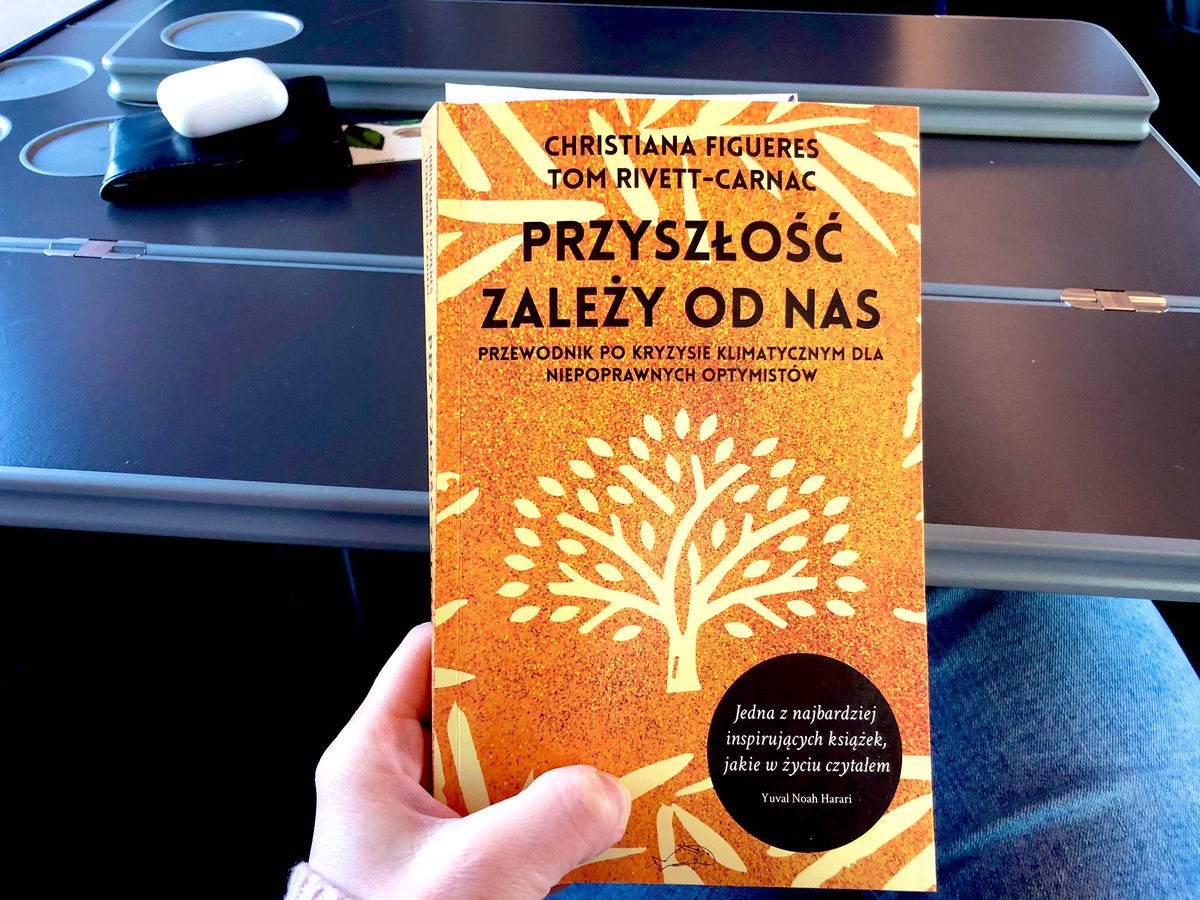 Zdjęcie z pociągu, widać rękę trzymającą książkę z pomarańczową okładka i drzewem - można odczytać autorów i tytuł z podtytułem: Christiana Figueres Tom Rivett-Carnac „Przyszłość zależy od nas. Przewodnik po kryzysie klimatycznym dla niepoprawnych optymistów”. W prawym dolnym rogu okładki czarne kółko z białymi literami: „jedna z najbardziej inspirujących książek, jakie w życiu czytałem” Yuval Noah Harari. W tle widać składany szary stolik w pociągu.