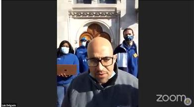 Screen shot Smart justice leader Luis Delgado testifying. Photo of other smart justice leaders in the background. 