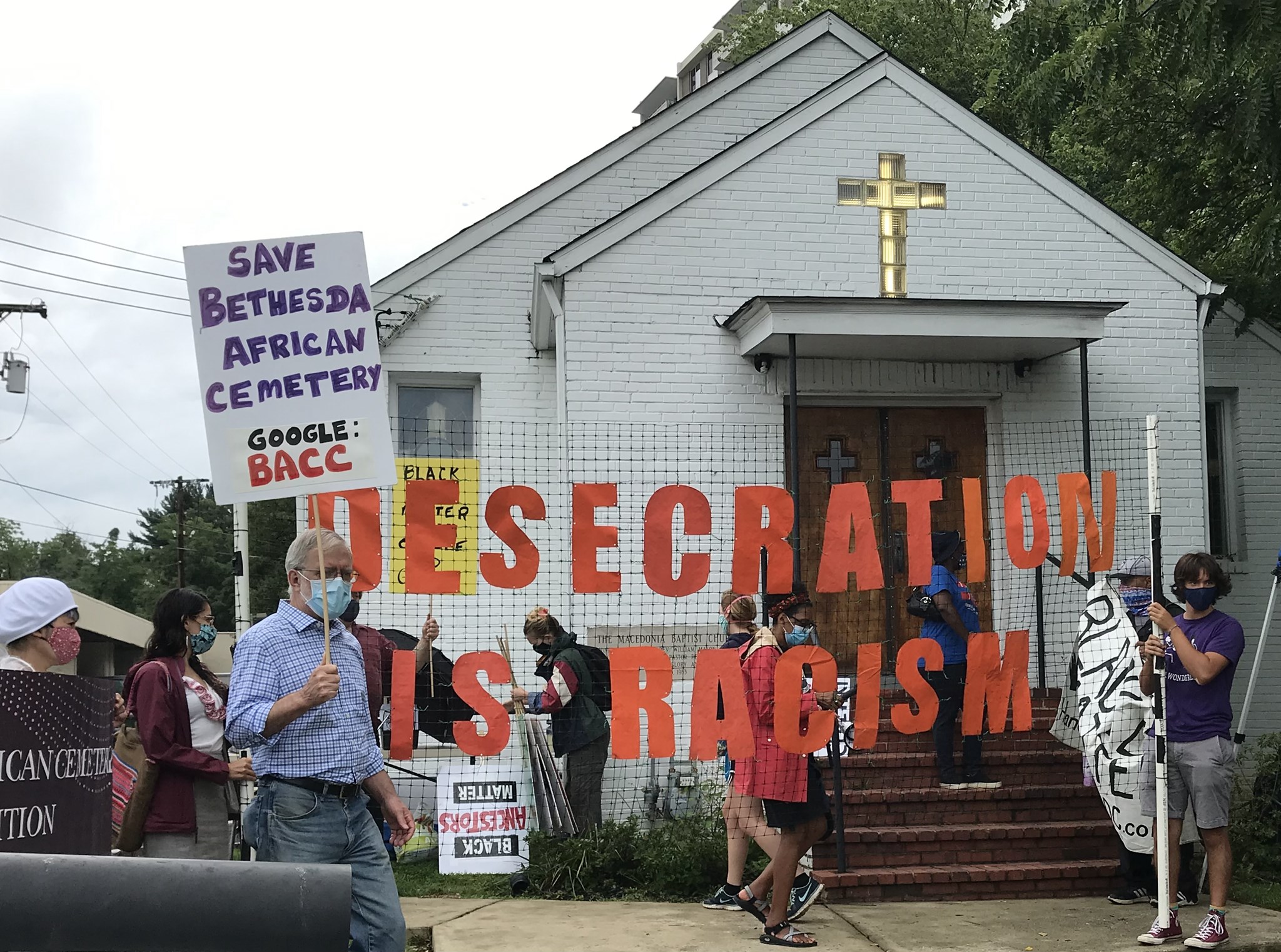 Multi-racial group of at least ten demonstrators on sidewalk outside of Macedonia Baptist Church, with "DESECRATION IS RACISM" sign