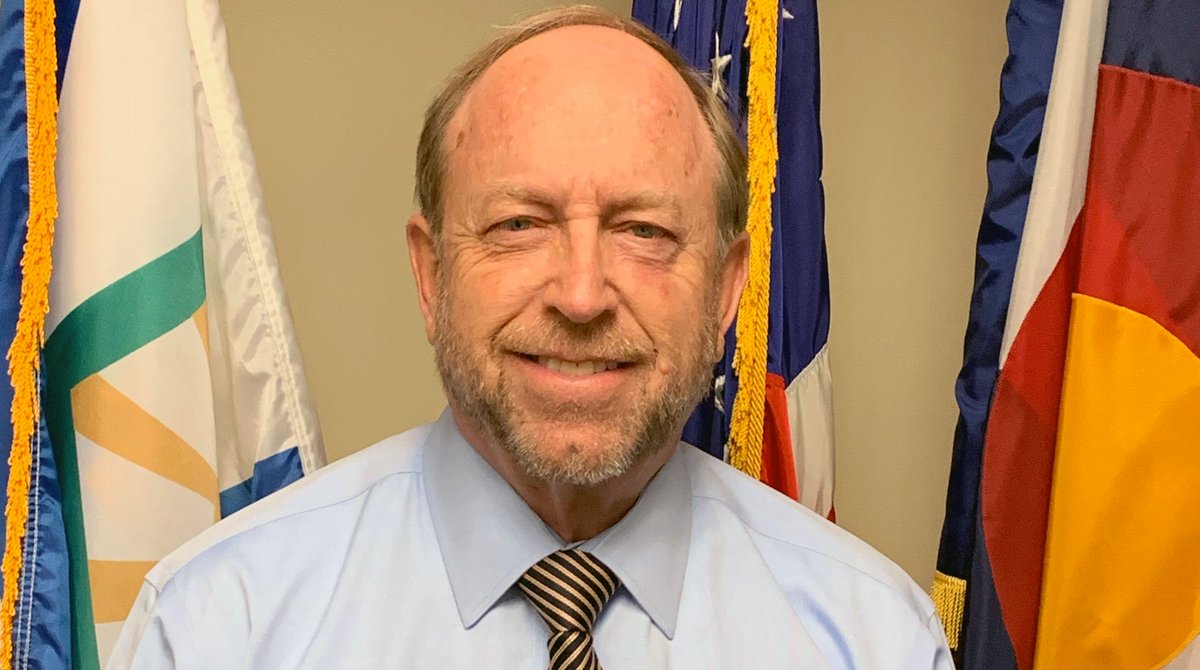 Mayor Suthers headshot, standing in front of three flags. His facial hair is scruffy.