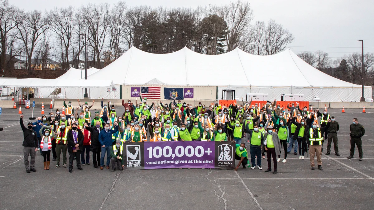 Dozens of workers and volunteers standing in front of the UAlbany vaccination site. They are proudly holding a banner that says 100,000+ vaccinations given at this site. 