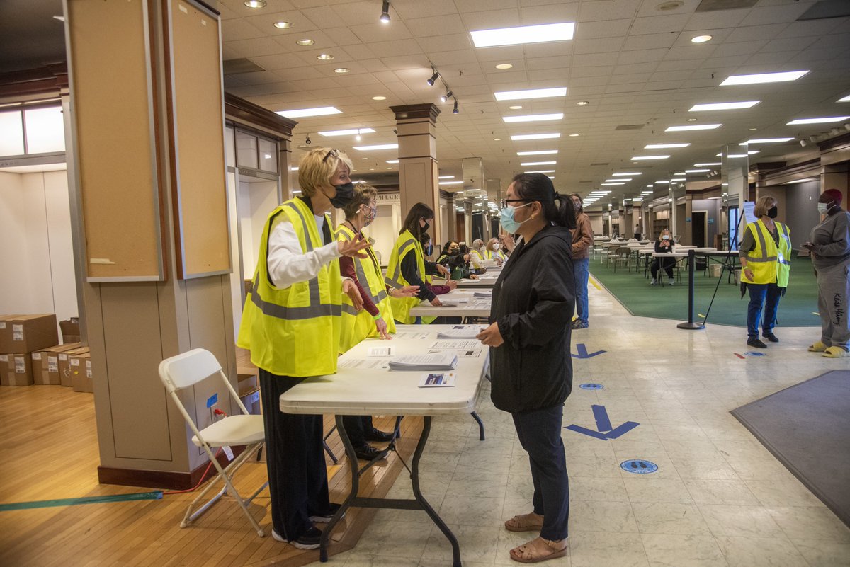woman in vest and mask speaking over a registration table to a vaccination client. there are arrows on the floor. there is a line of registration tables. people are waiting in line. 