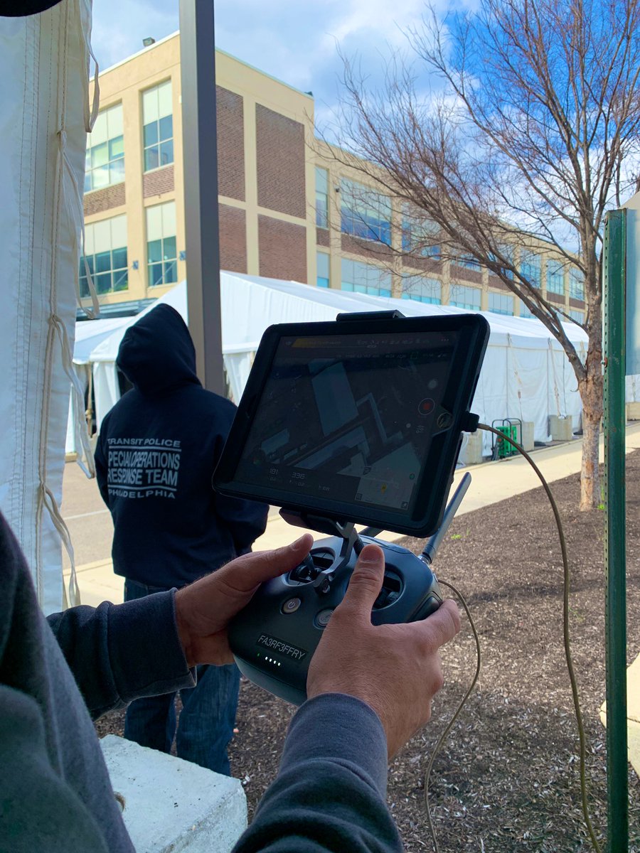Photo of a drone pilot with an aerial view camera. A person in the foreground has a hoodie that says transit police special operations response team Philadelphia. Tents and a building in the background.