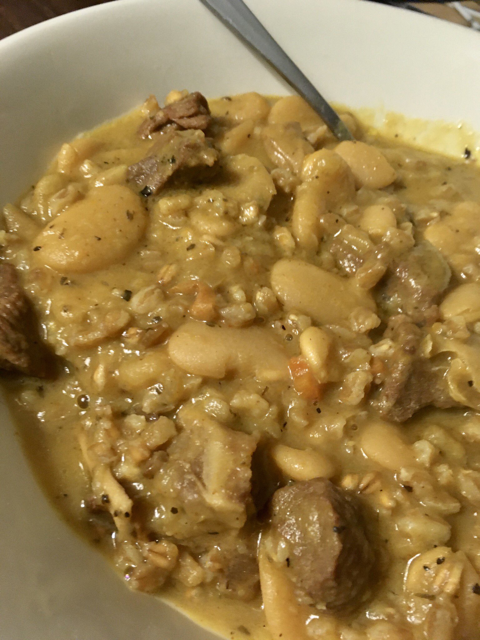 White bowl with spoon in it along with small pieces of goat, heirloom large white Lima beans from Rancho Gordo, garlic, onions, carrots, celery, half a chopped up squash, curry, Spanish Smoked Paprika, Cayenne, Ancho chili powder, farro, salt, pepper