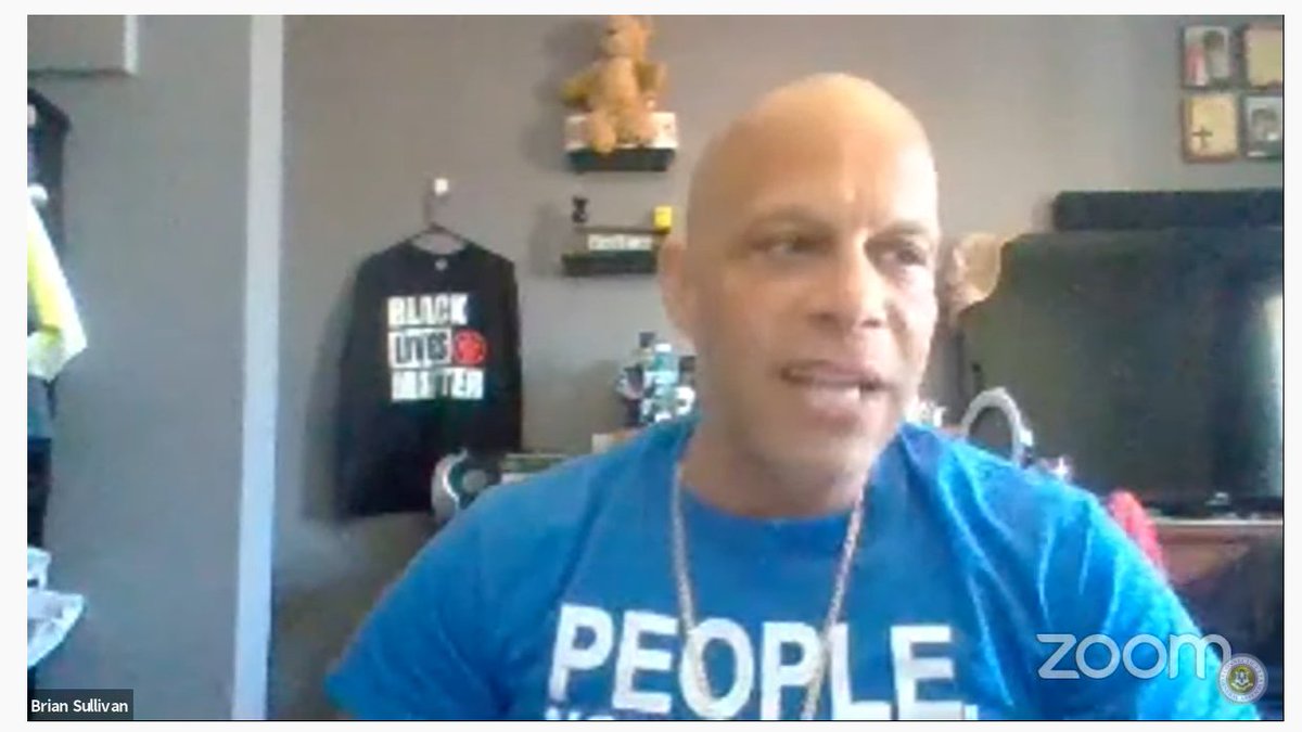 ACLU-CT Smart Justice leader Brian Sullivan testifies on Zoom before the Judiciary Committee. He is wearing a blue "people not prisons" Smart Justice shirt and is seating, looking slightly to the right. Behind him is a wall with shelves featuring a teddy bear and a Black Lives Matter shirt.