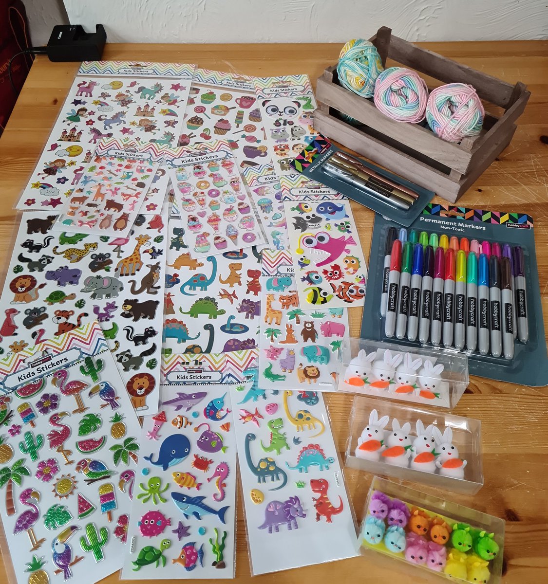 On the left, a large amount of colourful animal stickers on a table. On the right, from top to bottom: 3 balls of colourful yarn in a small wooden crate, a selection of colourful marker pens, little bunnies holding carrots, and colourful little Easter chicks