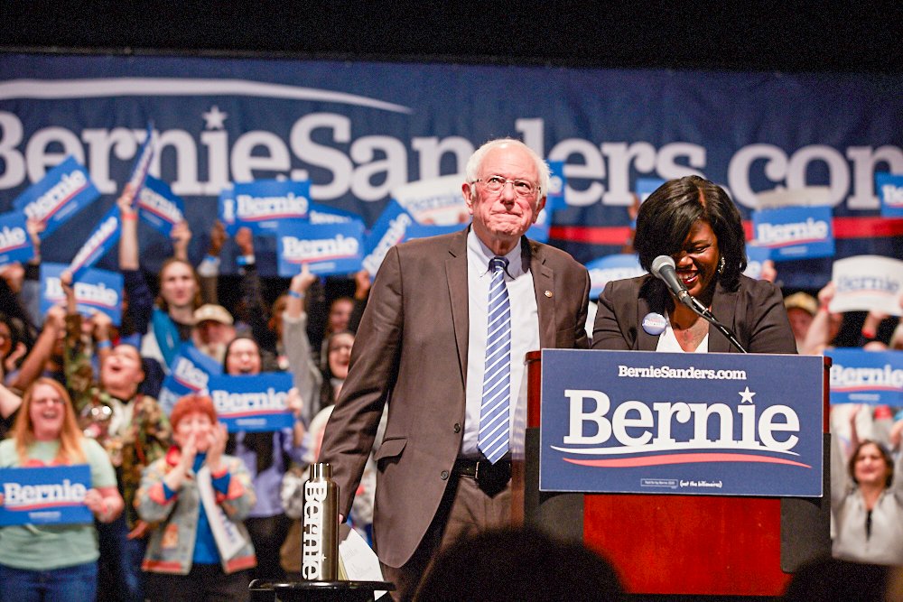 Cori Bush stands at a podium next to Bernie Sanders at a Bernie Sanders rally. 