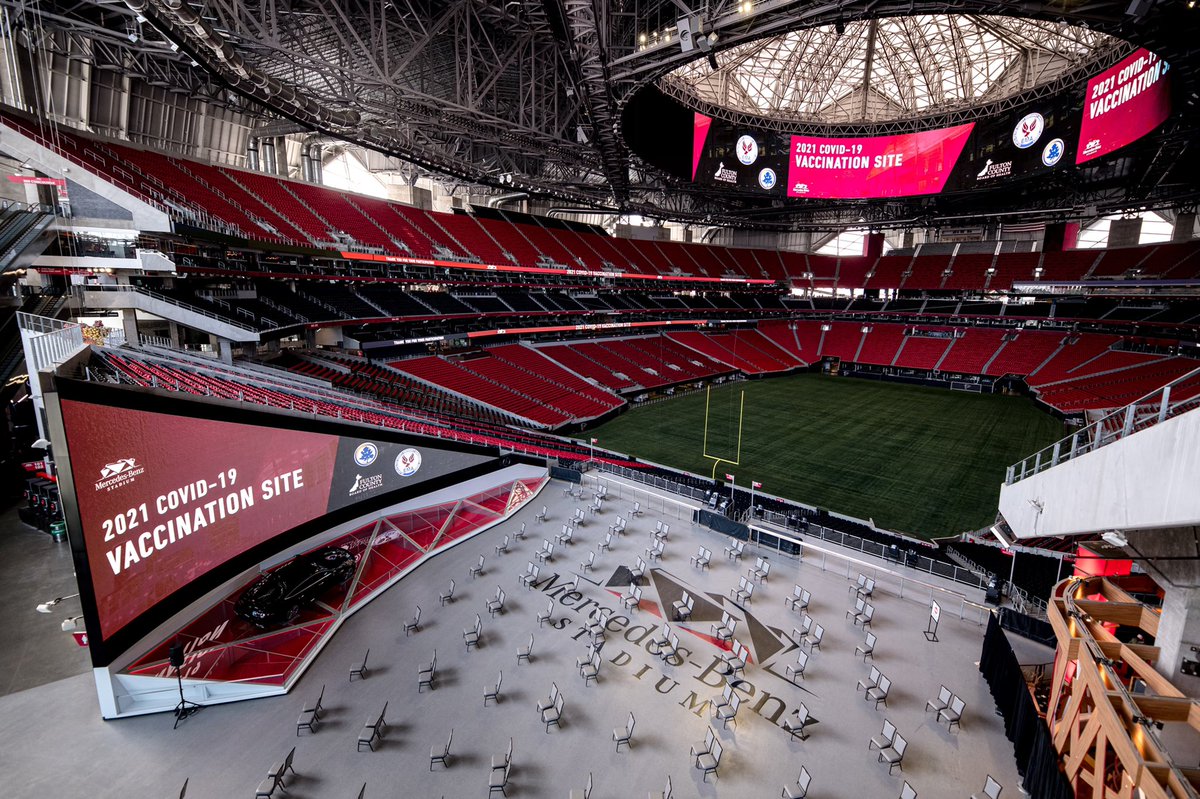 Overlooking Mercedes Benz Stadium. Seats are spaced out in groups of two. Field in distance. 2021 COVID-19 Vaccination Site signage displayed on side of stands.