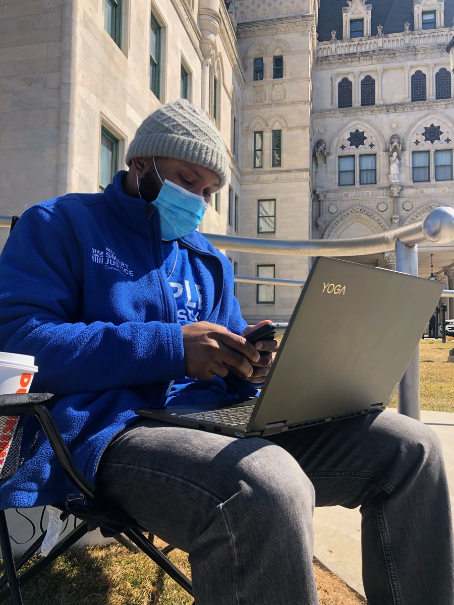 Smart Justice leader Timothy sitting on a chair in front of the capital with a laptop on his lap. He is wearing a blue Smart Justice fleece. He has a blue mask on and is on his phone as well. 