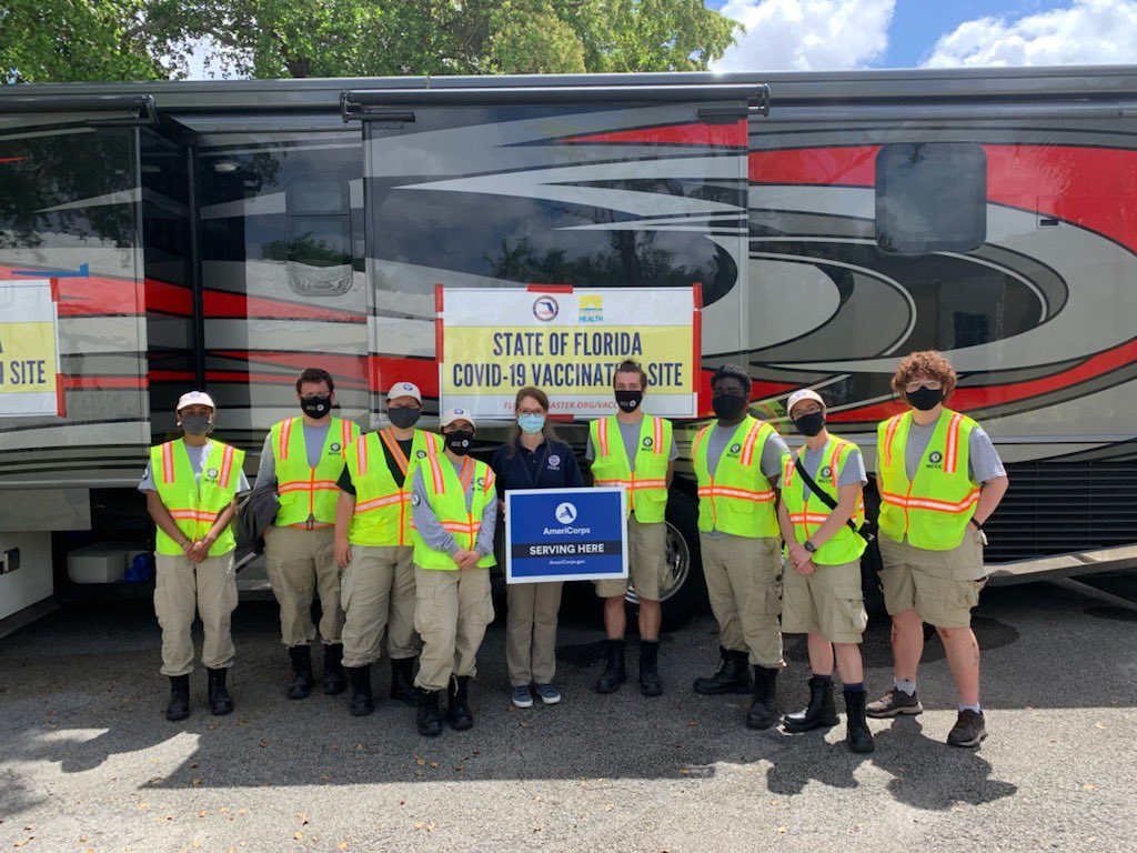 AmeriCorps members stand with Regional Administrator in front of large bus. Sign says serving here