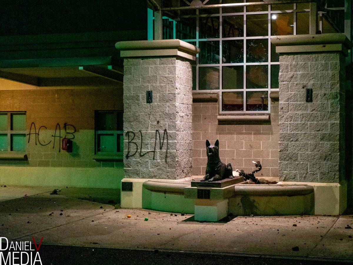 The front of the brick MCSO office. Windows at the front of the building are mostly shattered out, in front of them on a low wall is a bronze dog statue. There are two tags in black on the building reading "BLM" and "ACAB".
Light fixtures that are normally on this part of the building are missing, one lays on the wall next to the dog statue.