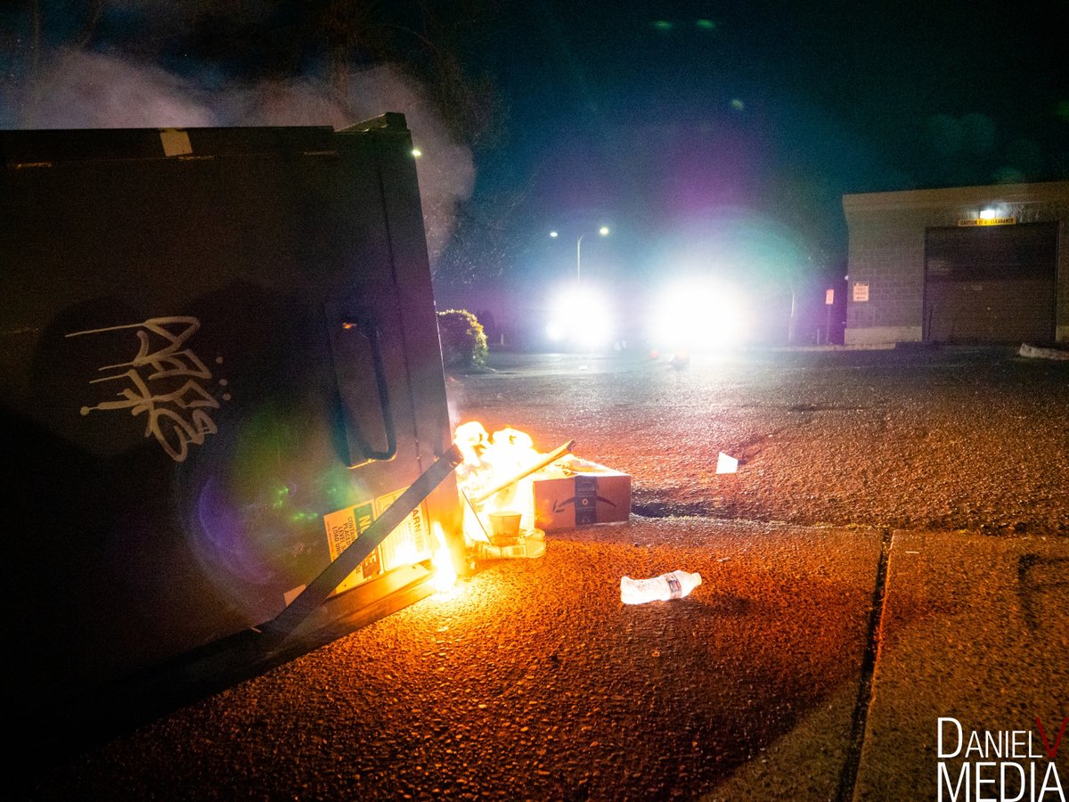 A dumpster is tipped over and on fire in the driveway of the MCSO Office. Two cops cars off in the distance on the side of the office are shining very bright lights directly into the camera.