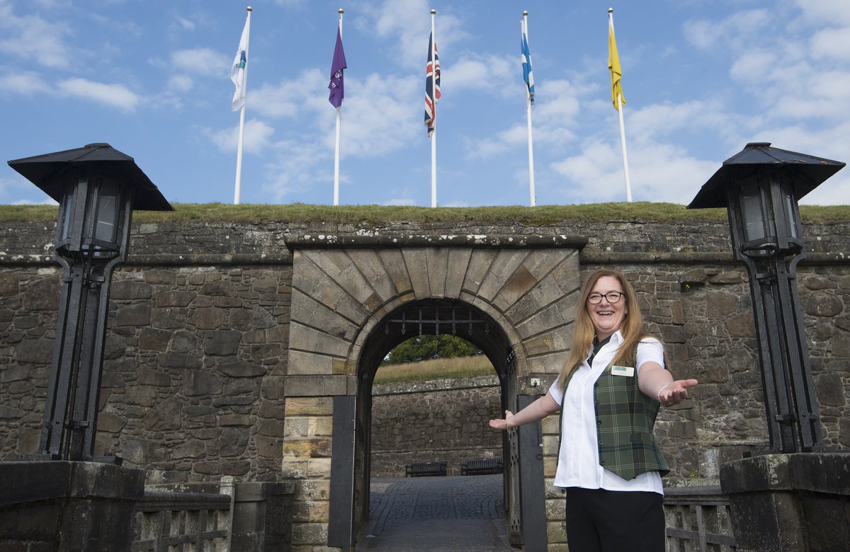 Woman in staff uniforms holding up her arms in a welcoming gesture in front of the castle gate at Stirling Castle. 