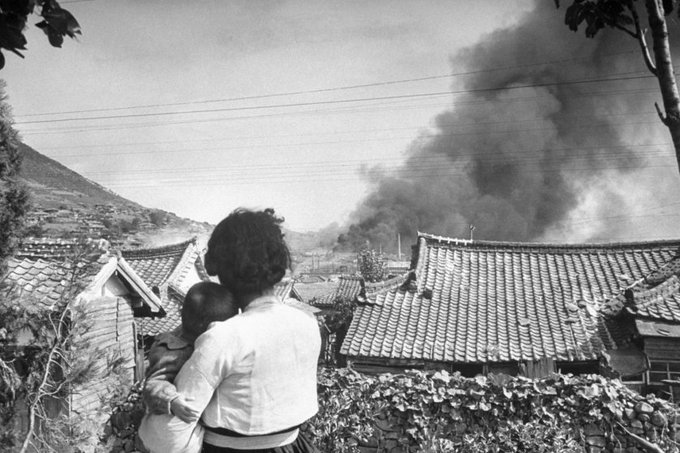 A woman holds a child close to her, facing away from the camera. In the distance a huge black plume of smoke is rising over the tiled roofs of a town.
