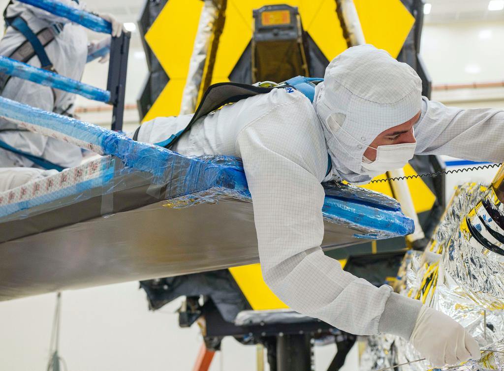 A technician hunches over a platform to carefully fold the sunshield layers of the James Webb Space Telescope.