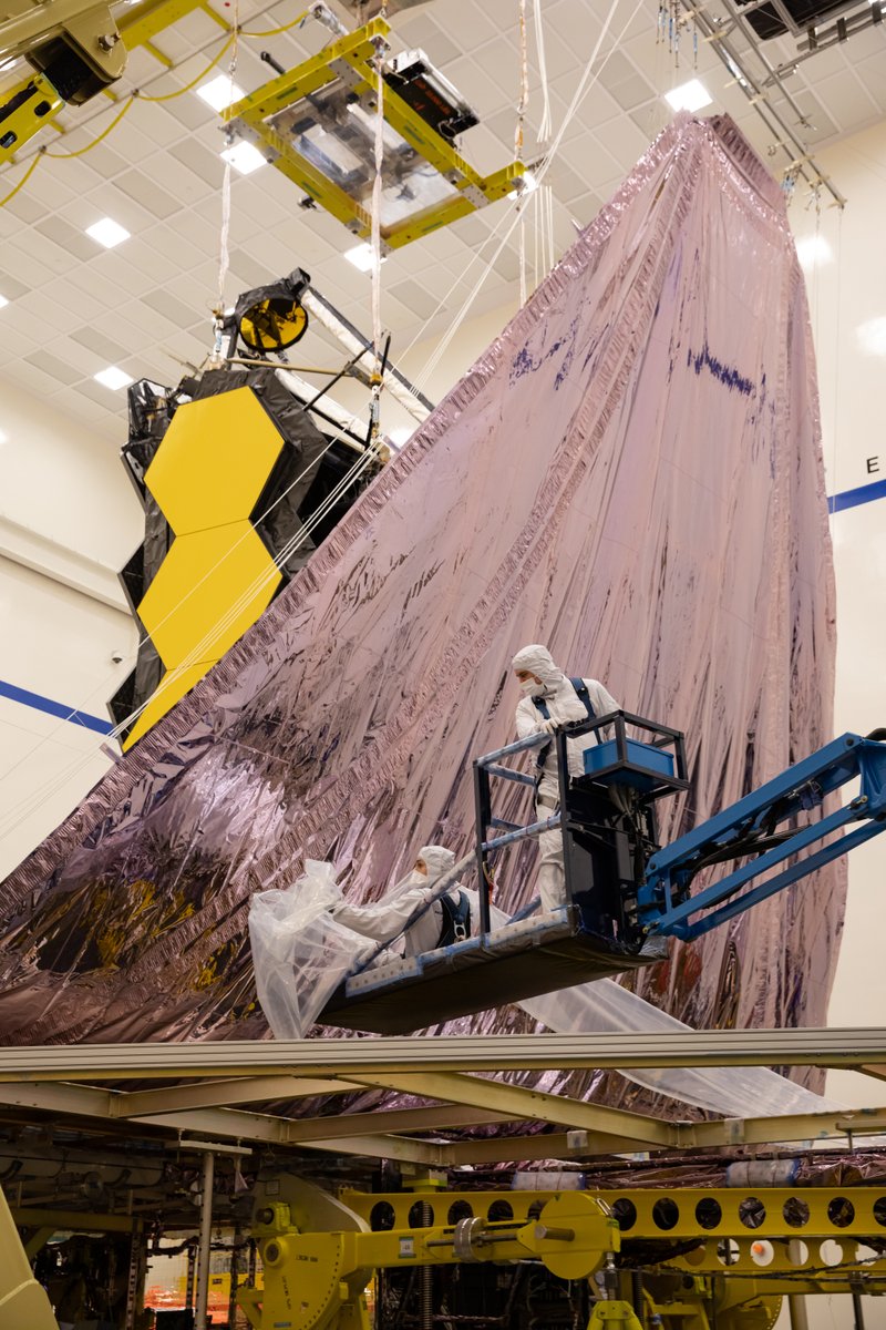 The sunshield of the James Webb Space Telescope resembles a sail as it is lifted vertically in preparation for sunshield folding. Two technicians stand on a crane on the right.