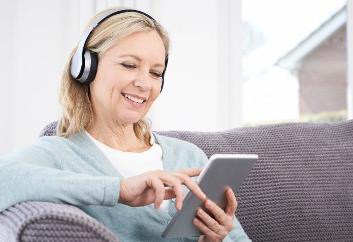 An older woman sits on a couch. She wears headphones and reads an audiobook on a tablet.