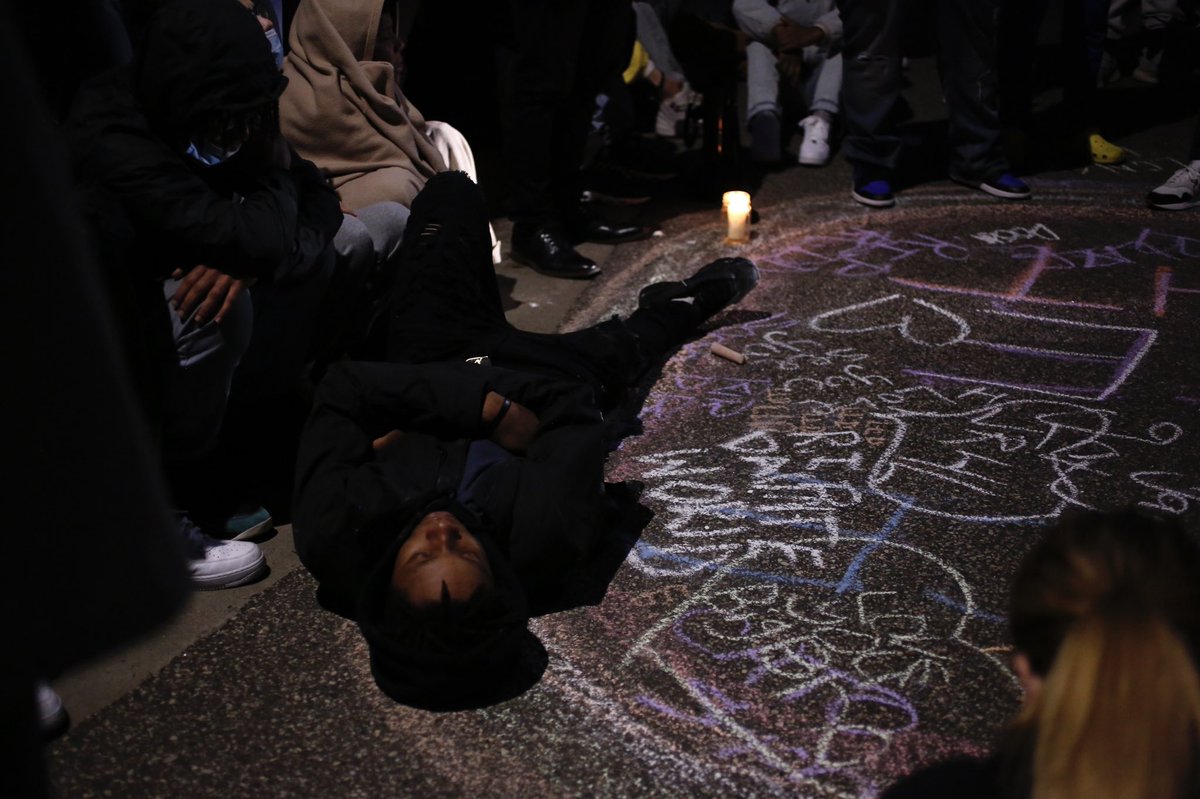 People continue to hold space at 63rd Ave and Lee Ave, circled around candles and a chalk memorial, with one man lying on the ground as a die-in