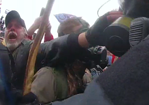 A man wearing a cap, dark-colored jacket, and a fingerless glove with a gray mustache and beard appears to be shouting while reaching out from within a crowd to grab someone's respirator.