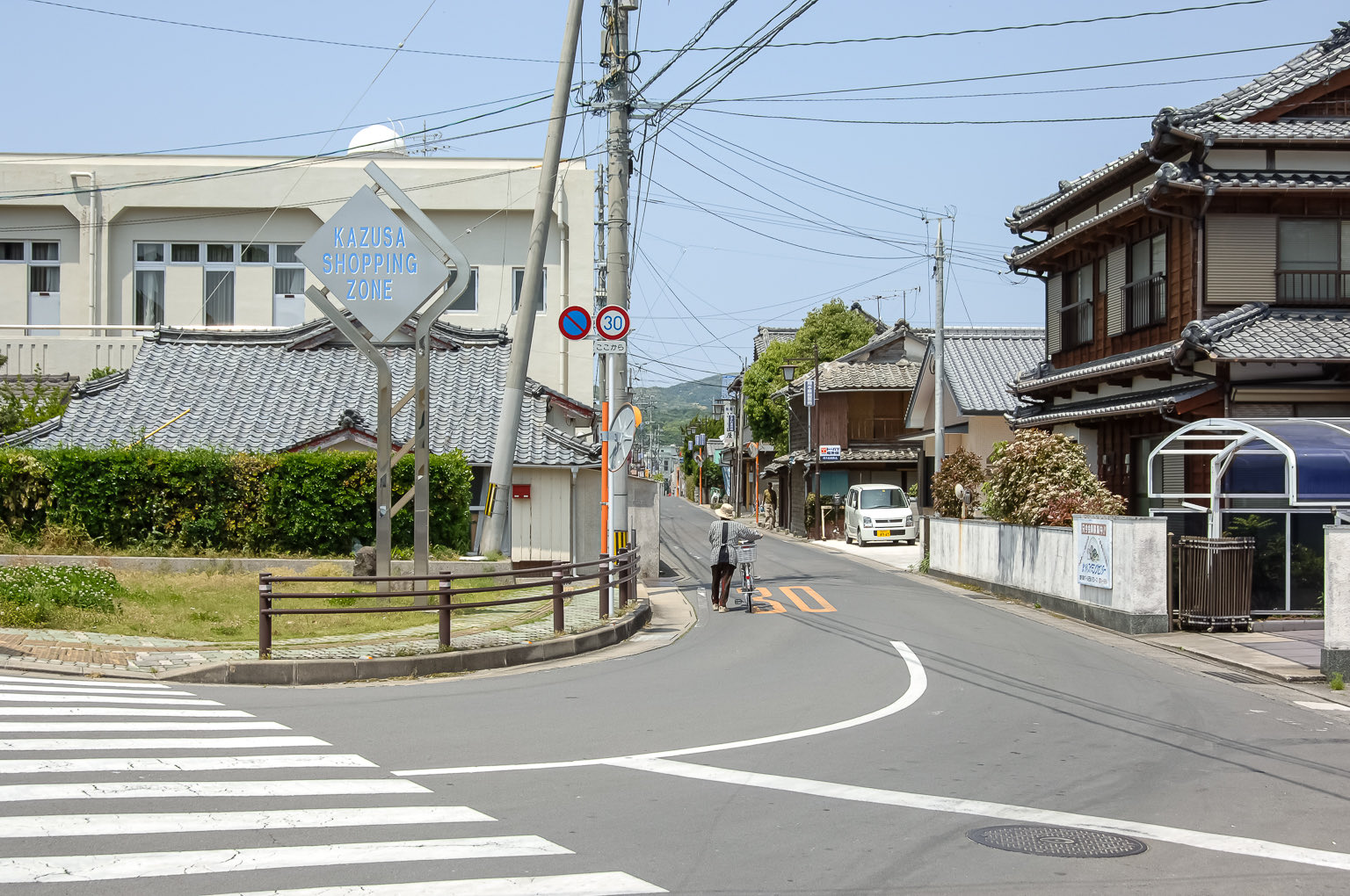 三日画師 南島原市加津佐町己 家並みの向こうに加津佐温泉神社 お四面さま が見える Kazusa Shopping Zone のシンボル塔が立つ加津佐駅前の三叉路に戻る 島原鉄道加津佐駅に停車する諫早行き2500形気動車 ここまで乗ってきた車両と同じである