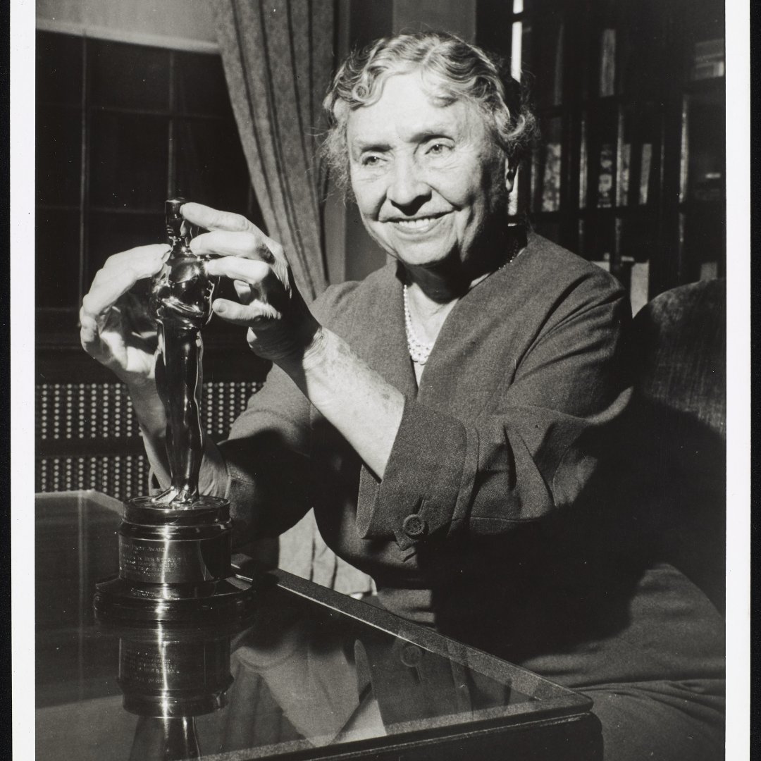 #ForYourConsideration: Helen Keller viewed from the waist up as she sits at a table at the American Foundation for the Blind. She is smiling and touching the Honorary Oscar that she received for the movie 'Helen Keller in Her Story' in 1955. #Oscars #HelenKellerArchive