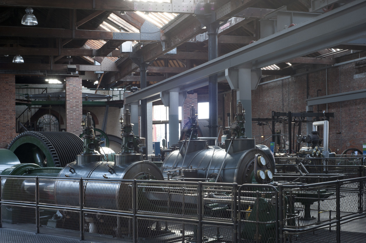 The old interior of the Power Hall at the Science and Industry Museum showing several standing steam engines that have large flywheels and boilers with brass gauges. The room is filled with steam.