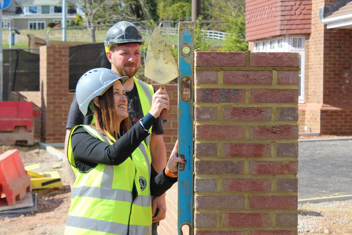 Jane Dodds holding a trowel and spirit level next to a newly built wall, having just laid a new brick.
