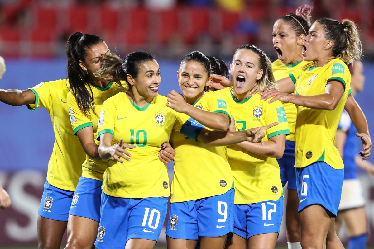 Jogadoras do Brasil, vestidas com a camisa amarela principal e calções azuis, comemoram juntas. Marta (com a 10), Debinha (com a 9) e Andressinha (com a 17), estão centralizadas. Ao fundo, a arquibancada desfocada. 