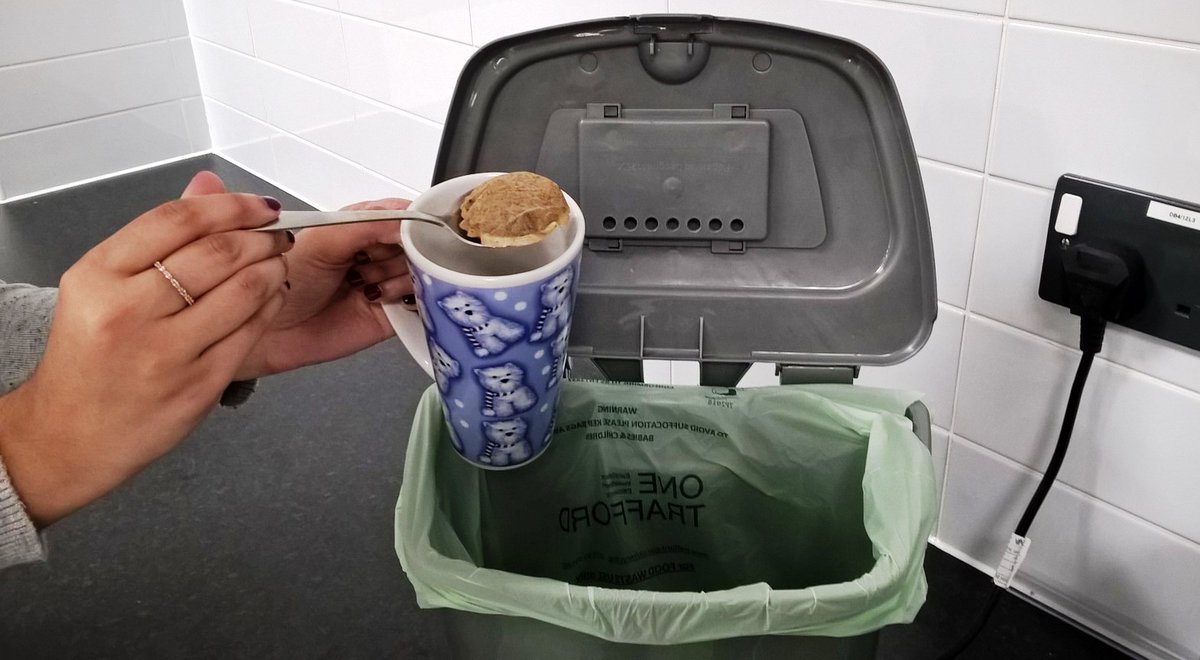 There is a food waste caddy on a kitchen counter. A person's hands are holding a mug and a spoon which is lifting a used tea bag out the mug. The person is hovering over mug and spoon over the caddy to place the tea bag in the recycling. 