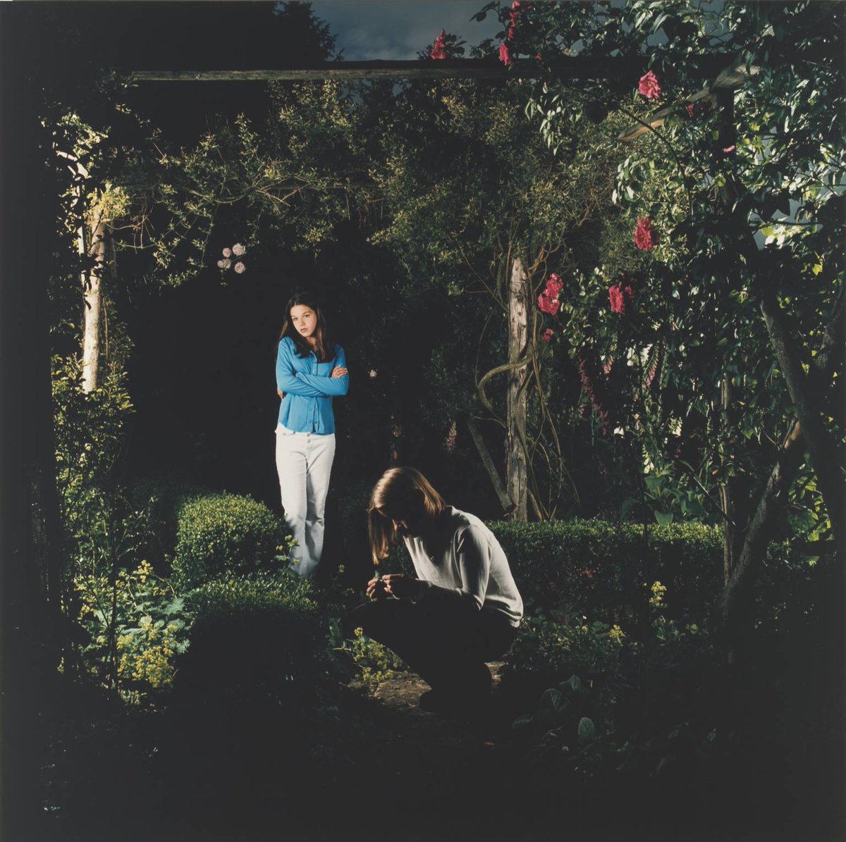 A colour photograph of two girls in an artificial looking garden. One girl is crouched and the other is stood nearby with her arms folded.